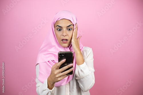 Young beautiful arab woman wearing islamic hijab over isolated pink background chatting, surprised and looking her phone © Irene