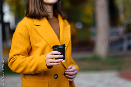 Female hand holding a cup of blank paper on a background of the park. Free space. Hands of a young girl in a yellow coat holding a paper cup of coffee. Girl hand hold coffee plastic cup. Coffee.