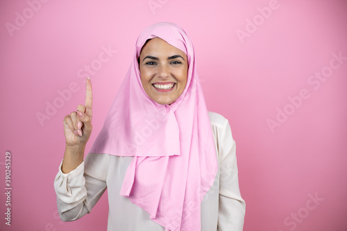 Young beautiful arab woman wearing islamic hijab over isolated pink background showing and pointing up with fingers number one while smiling confident and happy © Irene