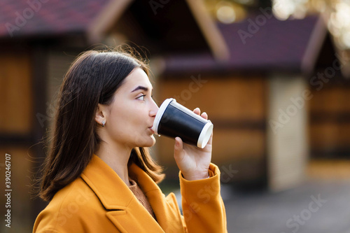 Female hand holding a cup of blank paper on the background of the park. Free space. The girl holds a cup of coffee and drinks coffee. Girl hand hold plastic coffee cup. Woman drinking coffee.