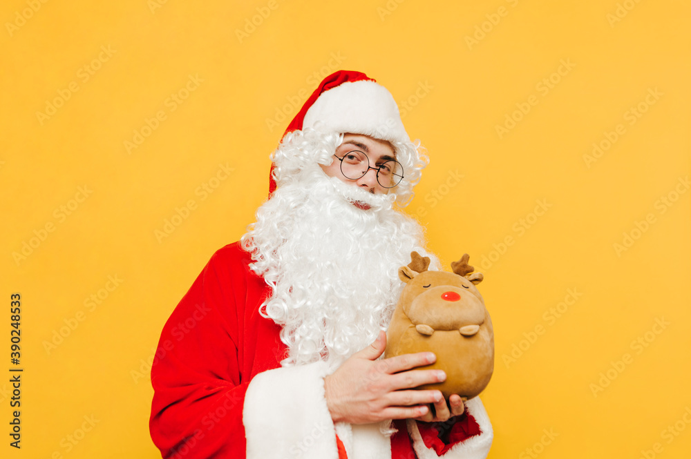 Cute Santa standing on a yellow background with a toy deer in his hands on a pose for the camera at Christmas. New year concept.