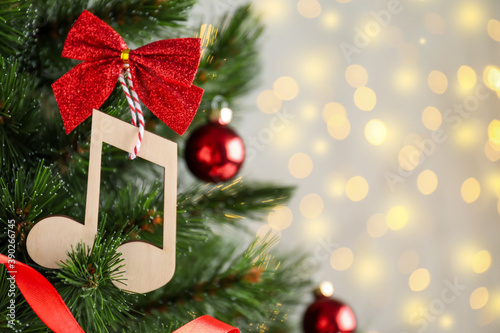 Wooden music note with red bow hanging on Christmas tree against blurred lights. Space for text