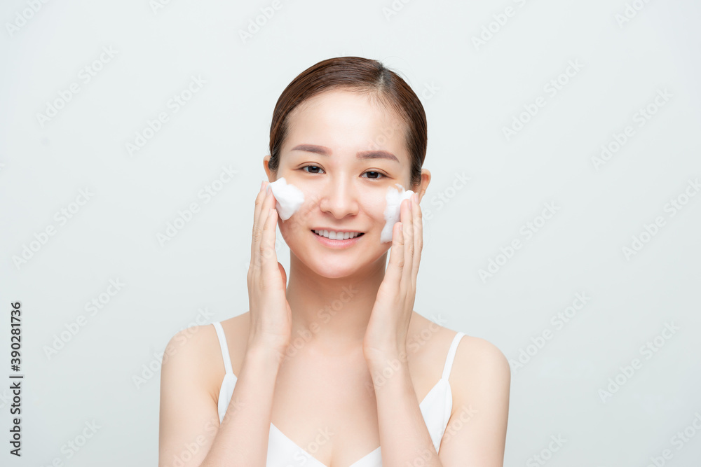 Satisfied beautiful woman makes massage of face, has white foam, cares of skin, smiles positively, models over white studio wall Satisfied beautiful woman makes massage of face, has white foam, cares of skin, smiles positively, models over white studio wall