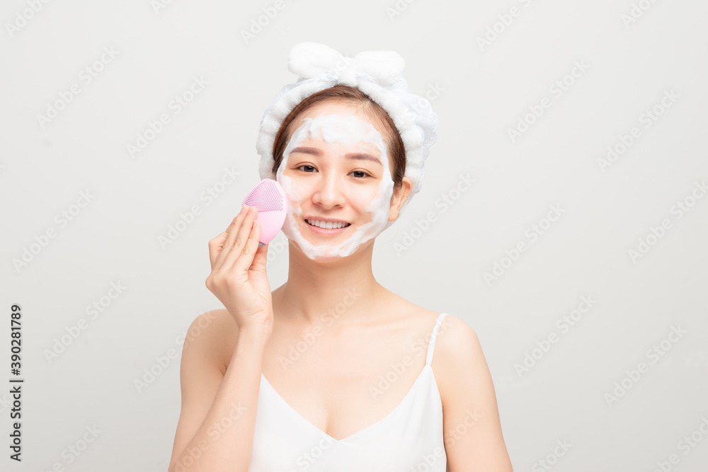 Young woman washing face with brush and cleansing foam on white background. Cosmetic products Young woman washing face with brush and cleansing foam on white background. Cosmetic products