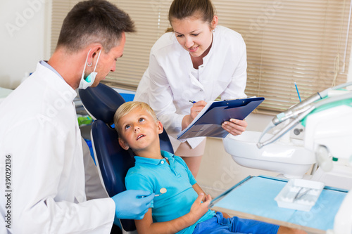 Dentist with assistant are diagnosticating to young patient which is sitting in dental clinic.