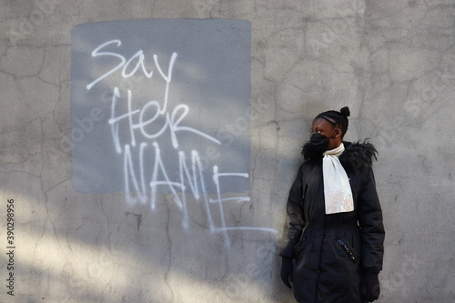 Student wearing mask standing looking at Say Her Name Spray painted words on cement wall