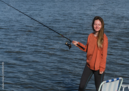happy girl with closed eyes stands on the shore of the lake with a fishing rod in hand and smiles