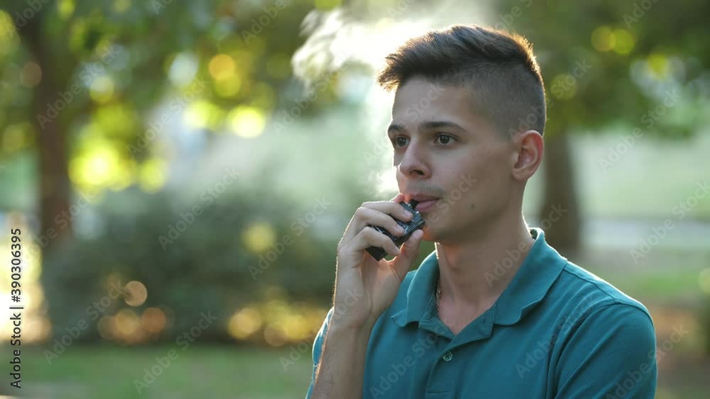 Friendly young man smoking e-cigarette in an airy green park area in summer Friendly young man smoking e-cigarette in an airy green park area in summer