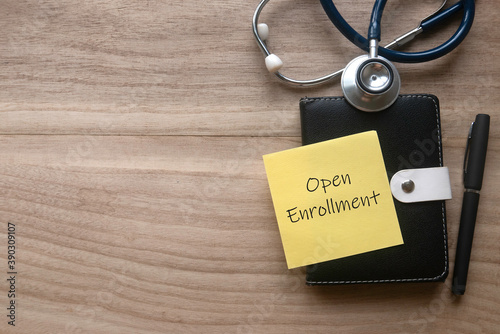 Top view of stethoscope, pen, notebook and yellow paper note written with Open Enrollment on wooden background with copy space.
