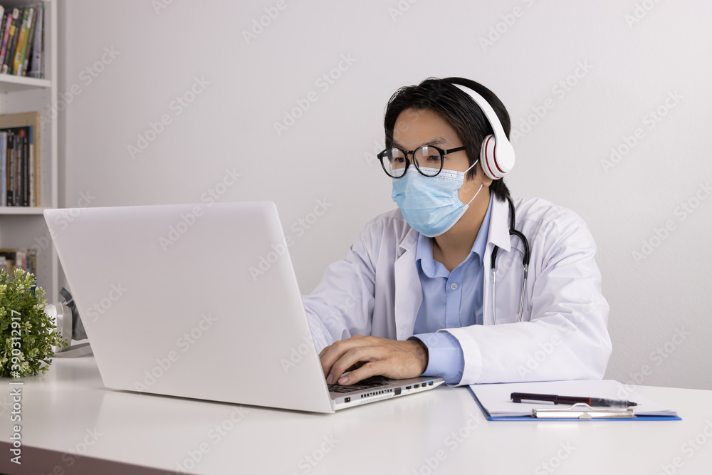 Young Asian Doctor Man in Lab Coat or Gown with Stethoscope Wear Face Mask and Glasses and Headphone Video Call or Video Conference with Patient Via Laptop Computer in Office Young Asian Doctor Man in Lab Coat or Gown with Stethoscope Wear Face Mask and Glasses and Headphone Video Call or Video Conference with Patient Via Laptop Computer in Office