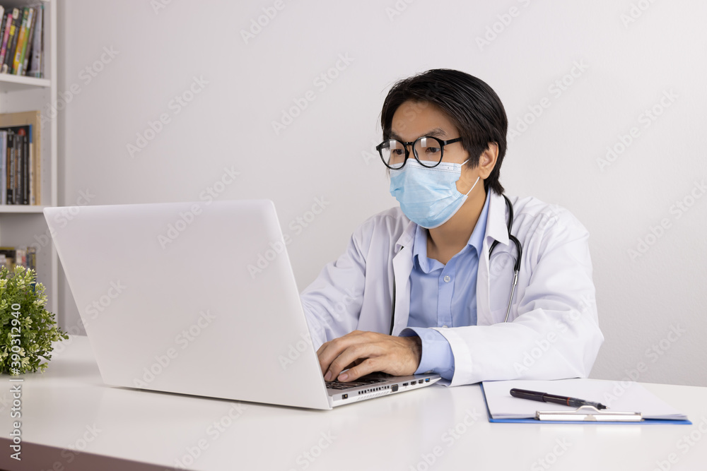 Young Asian Doctor Man in Lab Coat or Gown with Stethoscope Wear Face Mask and Glasses Using Laptop Computer on Doctor Table in Office Young Asian Doctor Man in Lab Coat or Gown with Stethoscope Wear Face Mask and Glasses Using Laptop Computer on Doctor Table in Office