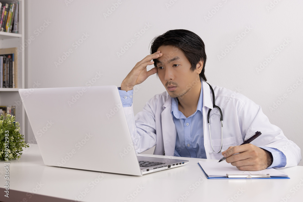 Serious Young Asian Doctor Man in Lab Coat or Gown with Stethoscope Writing Report and Using Laptop Computer on Doctor Table in Office Serious Young Asian Doctor Man in Lab Coat or Gown with Stethoscope Writing Report and Using Laptop Computer on Doctor Table in Office
