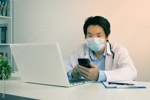 Young Asian Doctor Man in Lab Coat or Gown with Stethoscope Wear Face Mask Using Mobile Phone and Laptop Computer on Doctor Table in Office in Vintage Tone