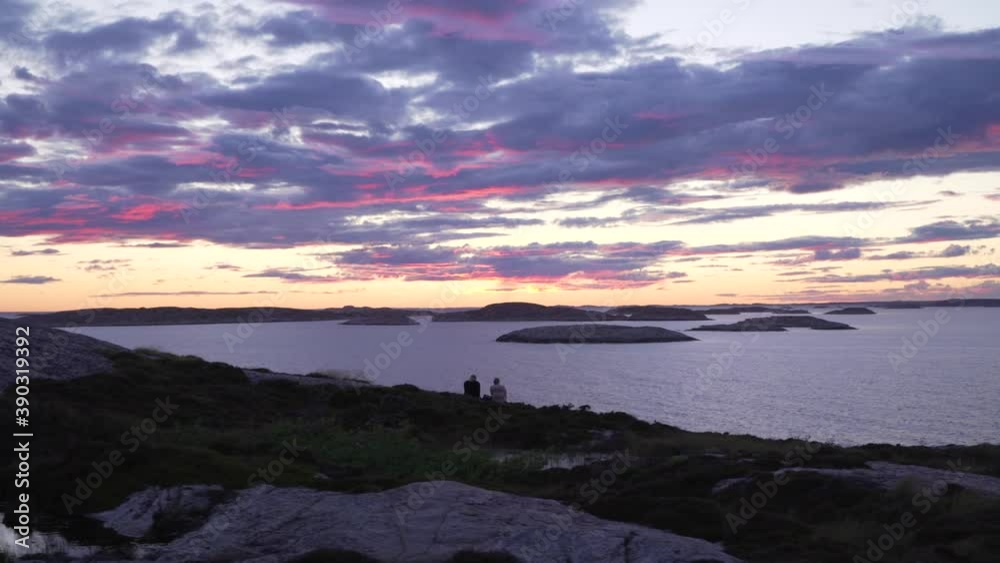 Couple watching sunset at ocean. Swedish coastline Bohuslan with islands and open sea with pink orange purple evening sky and clouds. Sweden west coast amazing unique archipelago landscape Couple watching sunset at ocean. Swedish coastline Bohuslan with islands and open sea with pink orange purple evening sky and clouds. Sweden west coast amazing unique archipelago landscape
