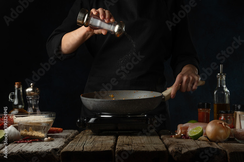 Professional chef pours salt into frying pan for cooking traditional indian curry. Backstage of preparing on dark blue background. 