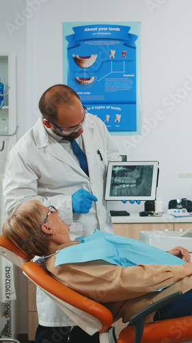 Dentist showing on tablet teeth x-ray reviewing it with patient. Doctor and nurse working together in modern stomatological clinic  explaining to old woman radiography of tooth using notebook display