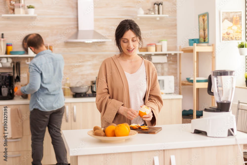 Woman peeling off oranges for nutritious smoothie in kitchen. Husband wearing denim shirt in the background. Healthy carefree and cheerful lifestyle, eating diet and preparing breakfast in cozy sunny