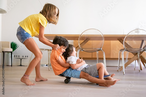 Cheerful children playing with skateboard at home. Blonde adorable girl pushing her two playful brothers. Happy kids riding on board and having fun. Childhood, game activity and weekend concept photo