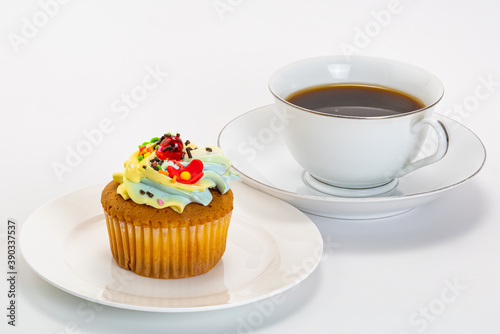 Vanilla cup cake garnished with butter cream frosting and colorful sprinkles in white ceramic plate and black coffee in white cup on white background with clipping path.