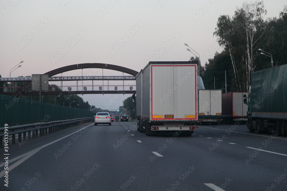European white van semi truck with trailer move on two lane suburban asphalted highway near metal bridge and trucks parking, back view at summer evening , road transportation cargo logistics