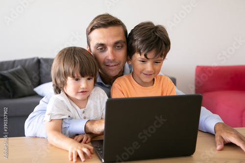 Happy middle-aged dad hugging sons and watching movie on laptop. Cute little boys sitting at table with father, looking at screen and smiling. Fatherhood, childhood and digital technology concept