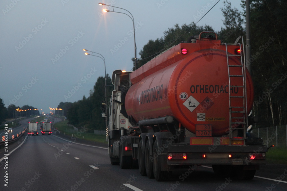 Modern orange semi truck fuel tanker with 30 1202 hazardous class sign and Russian inscription FLAMMABLE move on highway at summer evening in perspective, rear side view ADR DIESEL dangerous cargo