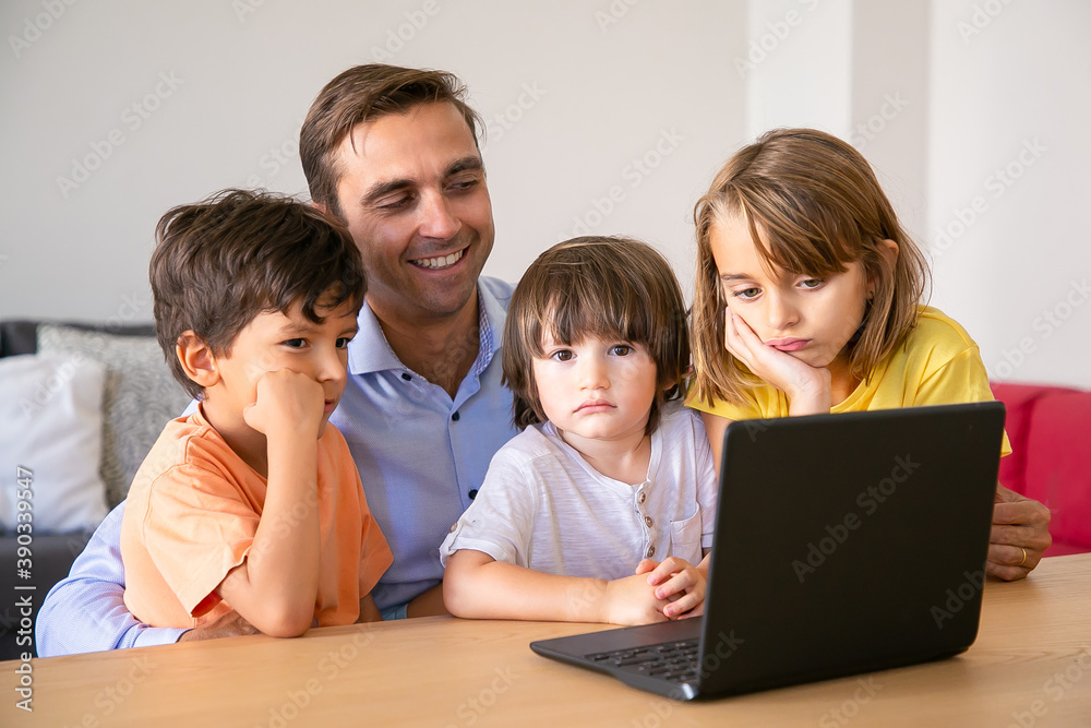Smiling dad and serious kids watching movie via laptop together. Middle-aged father sitting at table with children. Boys and girl looking at screen. Fatherhood and digital technology concept