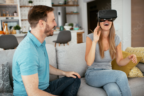 Smiling young woman using VR headset at home on couch. Woman and her husband enjoying virtual reality in apartment