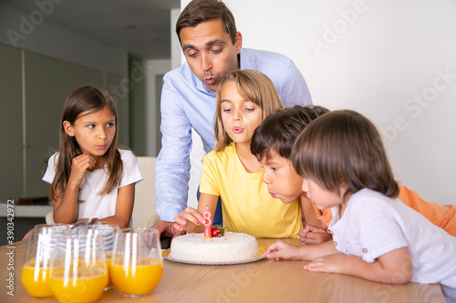 Happy kids and dad blowing out candle on cake and making wish. Pretty blonde girl celebrating her birthday with friends. Happy children standing near table. Childhood, celebration and holiday concept