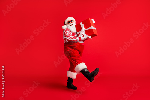 Full length photo of grandfather grey beard step hold big box white bow wear santa claus x-mas costume suspenders sunglass striped shirt cap boots isolated red color background