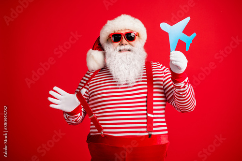 Photo of old man grey beard demonstrate blue paper plane funny wear santa claus x-mas costume suspenders sunglass striped shirt cap isolated red color background © deagreez