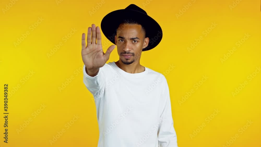 Portrait of serious hipster african american man showing rejecting gesture by stop palm sign. Guy isolated on yellow background.