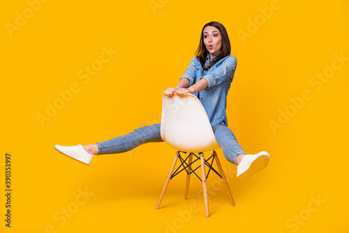 Full length photo of attractive pretty funny lady good mood sitting comfy chair spread legs hips fooling around wear casual denim shirt white shoes isolated bright yellow color background
