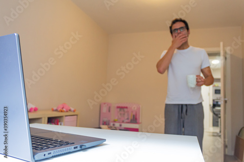 businessman working at home reaching his table with laptop, coffee in hand and yawning because he is just awake