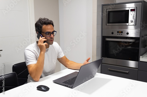 businessman working at home from kitchen, talking on mobile phone and looking at his laptop screen.