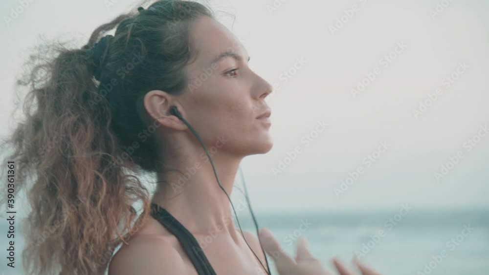 Close-up profile of young sun-tanned Caucasian woman wearing wired earphones stretching arms and neck before exercise, standing on ocean coast Close-up profile of young sun-tanned Caucasian woman wearing wired earphones stretching arms and neck before exercise, standing on ocean coast