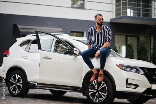 Successful arab man wear in striped shirt and sunglasses sitting on the hood of white suv car. Stylish arabian men in transport. © AS Photo Family