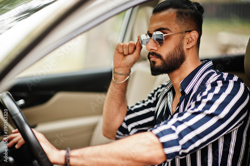 Successful arab man wear in striped shirt and sunglasses pose behind the wheel of  his white suv car. Stylish arabian men in transport.