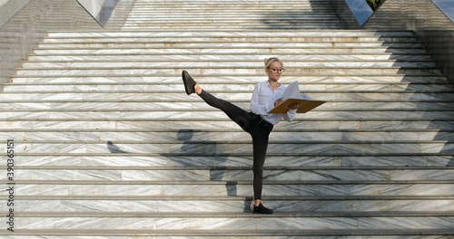Flexibility in a work with papers and documents. A beautiful girl, standing on the street stairs, revises documents in a yellow folder, lifts her right leg up.