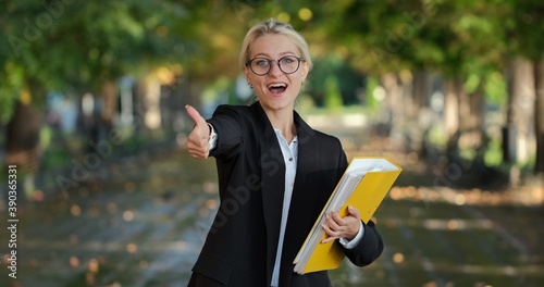 A beautiful girl in glasses stands with a yellow folder in the street in the center of the frame and shows her thumb up.
