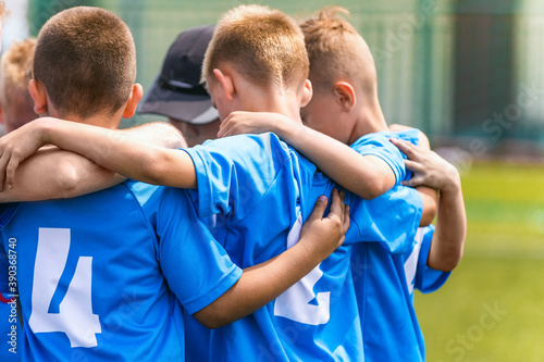 Kids soccer team with coach in group huddle before the match. Elementary age children are listening together to coach motivational speech. Boys in blue football shirts