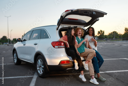 A group of three young well-dressed women of different nationalities sitting in an opened car trunk outside on a parking site smiling and laughing speaking to each other © Svitlana