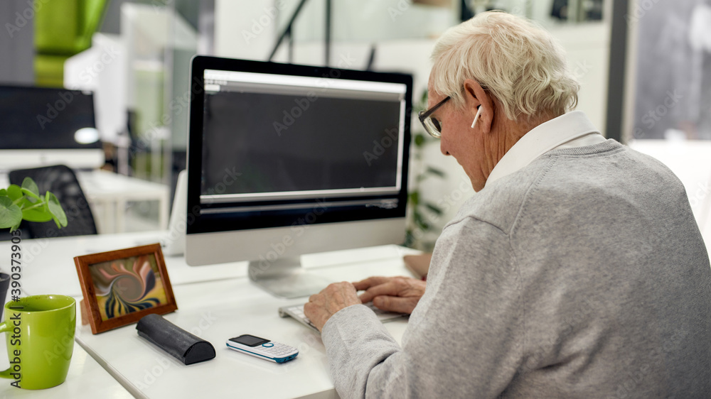 Back view of aged man, senior intern in wireless earphones looking focused while typing, using pc, sitting at desk, working in modern office Back view of aged man, senior intern in wireless earphones looking focused while typing, using pc, sitting at desk, working in modern office