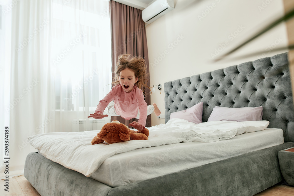 A little barefoot girl enjoying jumping down on her teddybear with her hair back on a bed A little barefoot girl enjoying jumping down on her teddybear with her hair back on a bed