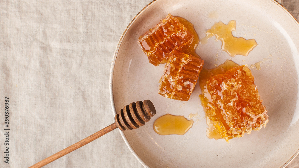 Fototapeta premium Flatlay honey in honeycomb on a plate with a wooden honey spoon on a linen tablecloth. Copy space.