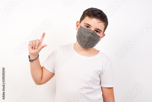 Caucasian young boy wearing medical mask standing against white background smiling and gesturing with hand small size, measure symbol.
