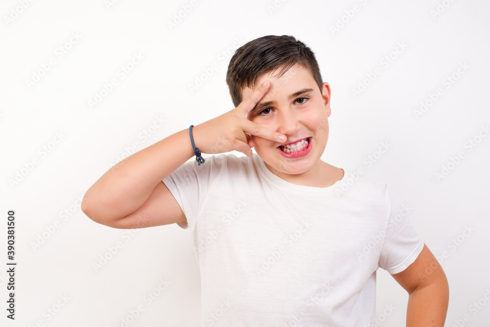 Caucasian young boy standing against white background making v-sign near eyes. Leisure, coquettish, celebration, and flirt concept.