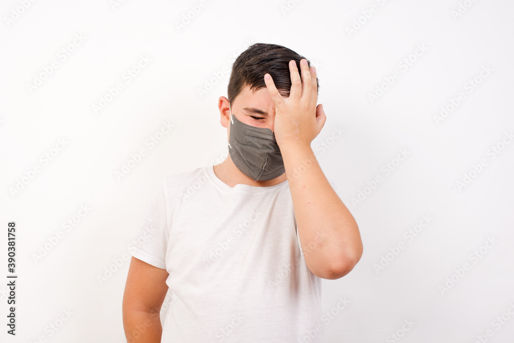Frustrated Caucasian young boy wearing medical mask standing against white background holding hand on forehead being depressed regretting what he did having headache, looking stressful. Frustrated Caucasian young boy wearing medical mask standing against white background holding hand on forehead being depressed regretting what he did having headache, looking stressful.