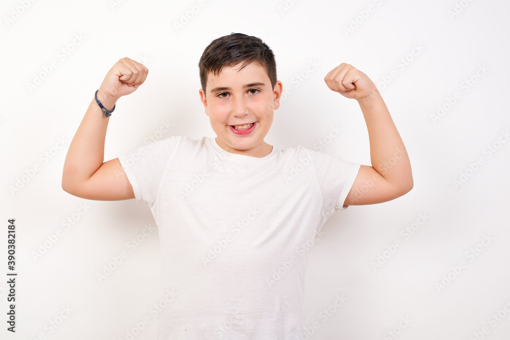 Waist up shot of Caucasian young boy standing against white background raises arms to show muscles feels confident in victory, looks strong and independent, smiles positively at camera. Sport concept. Waist up shot of Caucasian young boy standing against white background raises arms to show muscles feels confident in victory, looks strong and independent, smiles positively at camera. Sport concept.