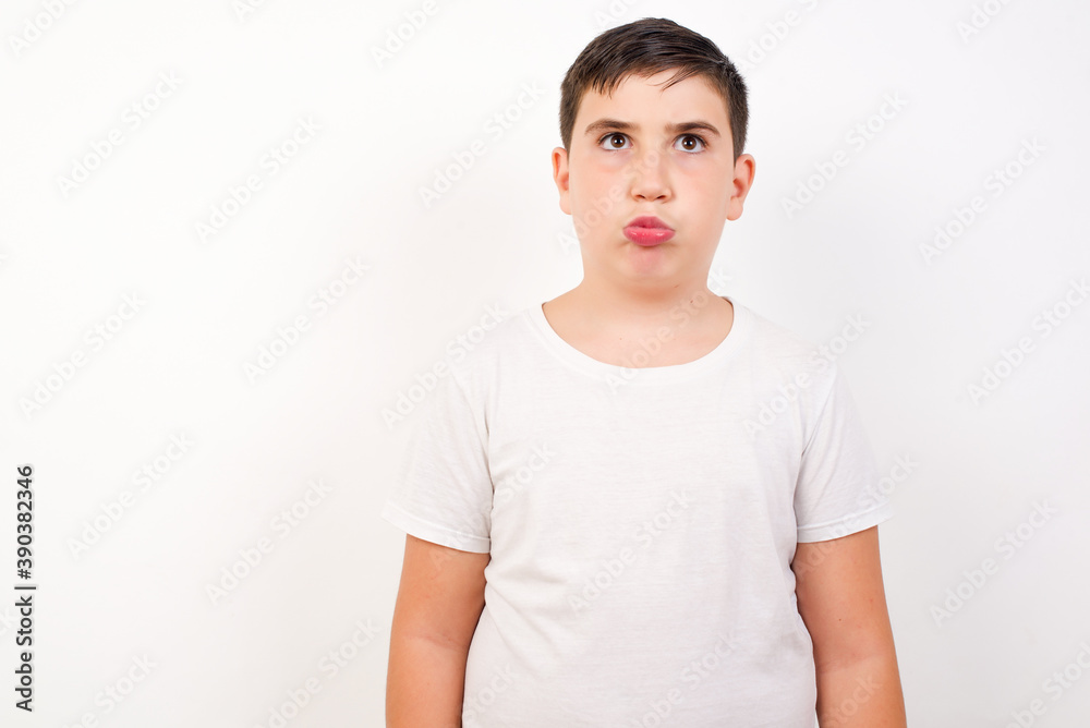 Gloomy, bored Caucasian young boy standing against white background  frowns face looking up, being upset with so much talking hands down, feels tired and wants to leave.
